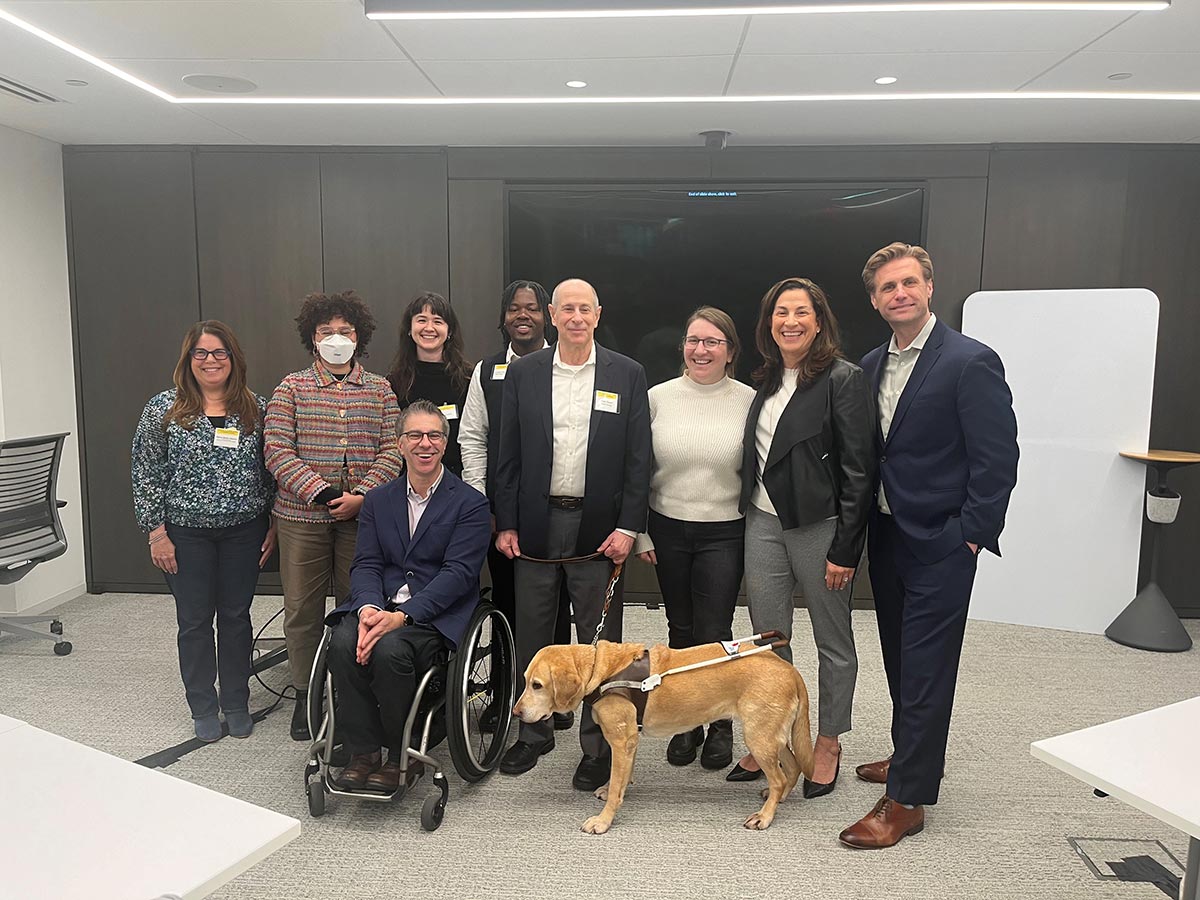 Peter with his guide dog Inga, a yellow lab guide dog is at the center on a harness, standing next to a man in a wheelchair. Everyone is smiling, wearing business attire with name badges, in front of a large wall-mounted screen and wood-paneled wall.